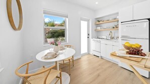 A modern kitchen with white cabinets, a built-in oven, and a refrigerator. There is a dining table with chairs and a window with a view of greenery outside. - Cute Retro Studio King Bed & Workspace - Villa 10 (Lake Worth)