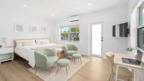 A modern hotel room with a bed, two armchairs, a desk with a laptop, a TV, and a window with a view of greenery. - Cute Retro Studio King Bed & Workspace - Villa 10 (Lake Worth)