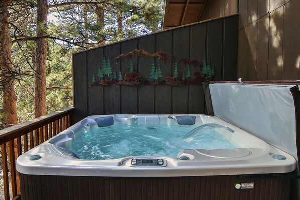 A hot tub with a control panel, surrounded by a wooden deck and a forest backdrop.