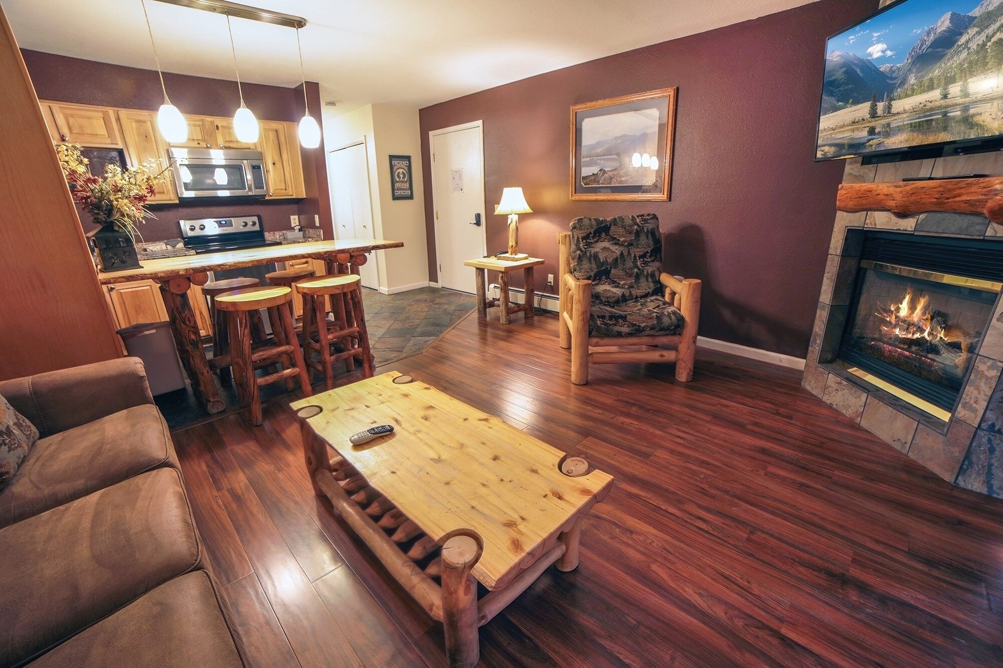 A cozy living room with a fireplace, wooden furniture, and a kitchen area with wooden cabinets and a stove.