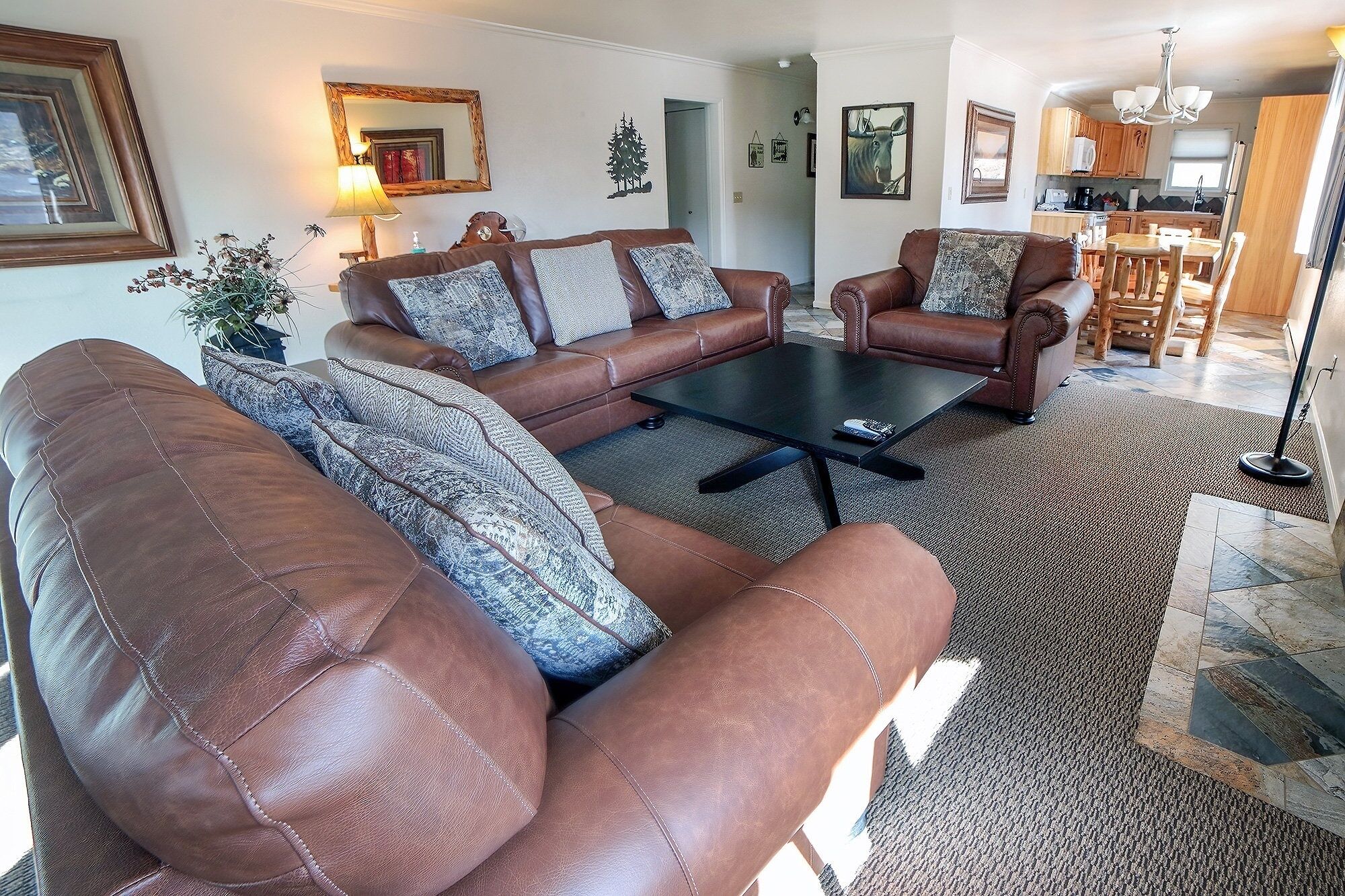 A living room with a brown leather sofa, a matching armchair, and a coffee table. There is a rug on the floor, and a kitchen is visible in the background.