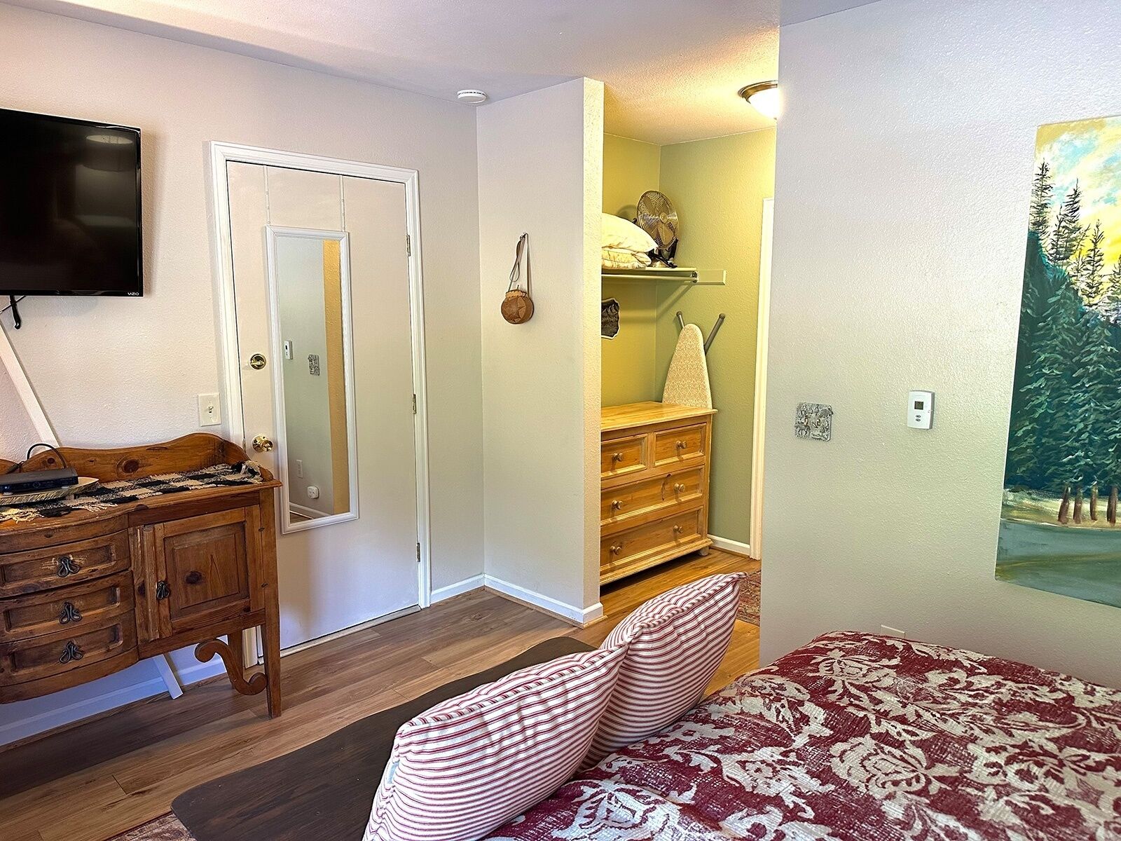 Una stanza con una cassettiera in legno, un letto con biancheria a motivi decorativi, un televisore montato a parete e un quadro raffigurante una foresta.