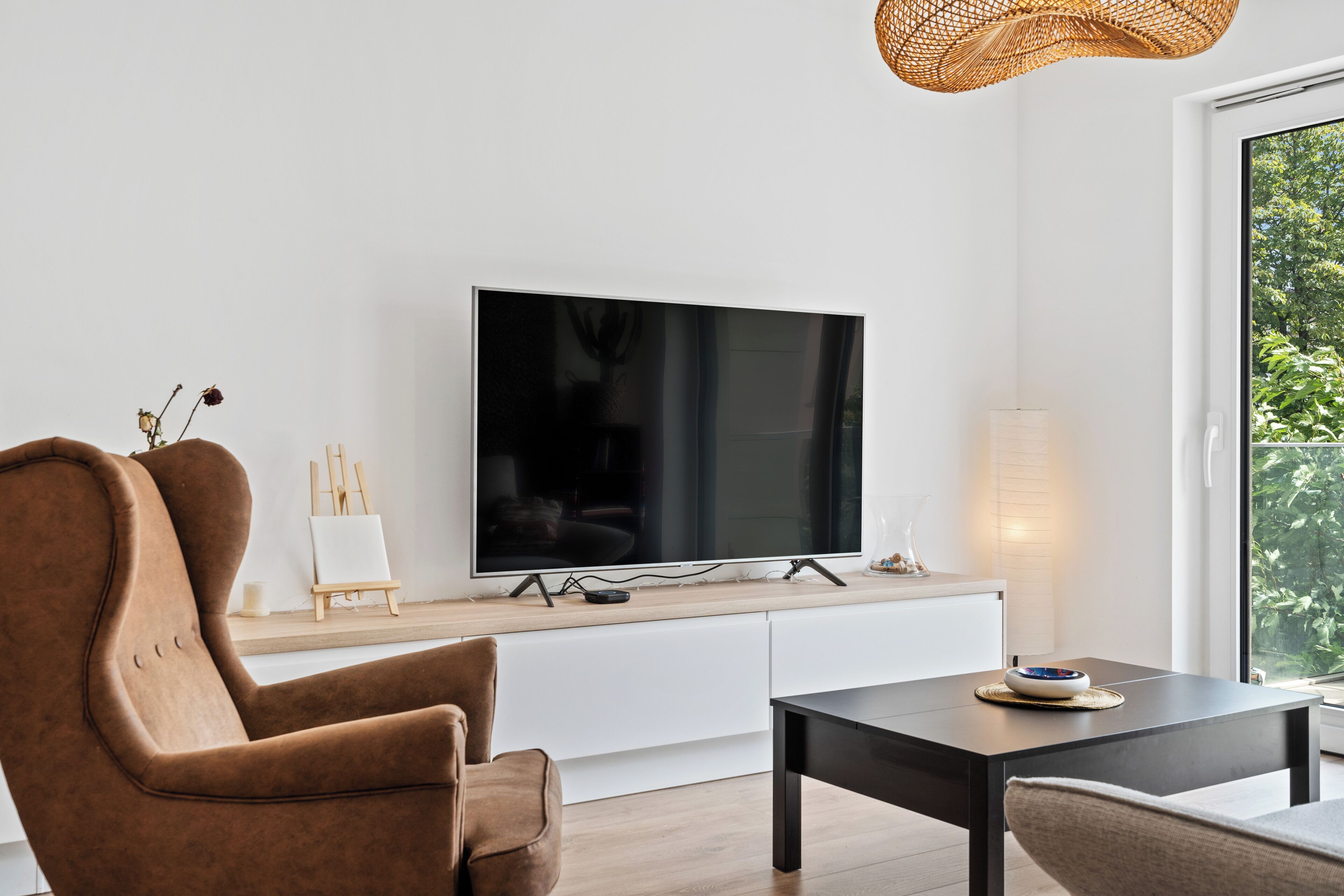 Ein modernes Wohnzimmer mit Flachbildfernseher an einer weißen Wand, einem braunen Ledersessel, einem schwarzen Couchtisch und einer Weiden-Pendelleuchte.