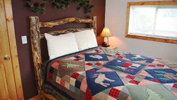 Quarto com cabeceira de madeira rústica, colcha acolchoada, mesa de cabeceira com abajur e janela com persianas.