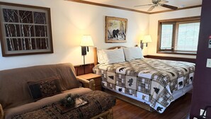A bedroom with a bed, a sofa, a nightstand, a lamp, a ceiling fan, and a window with blinds.