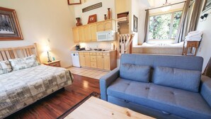 A cozy room with a bed, a blue sofa, a wooden coffee table, and a kitchen area with cabinets and a microwave. - Sunnyside Knoll: 18 1 Bedroom Cabin (Estes Park)
