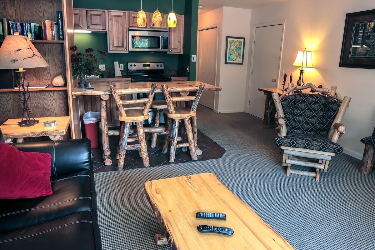 Een gezellige woonkamer met een houten salontafel, een leren bank en een fauteuil met een patroon. In de achtergrond is een keuken met houten keukenkastjes en een magnetron zichtbaar.