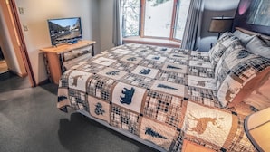 Un dormitorio con una cama grande, un televisor sobre una mesita de noche de madera, una ventana con cortinas y una lámpara en la mesita.