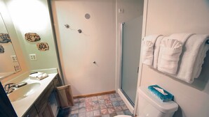 A bathroom with a white sink, a mirror, a toilet, and a shower. There are towels hanging on the wall and a box of toilet paper on the toilet. - Sunnyside Knoll: 12 1 Bedroom Apts (Estes Park)