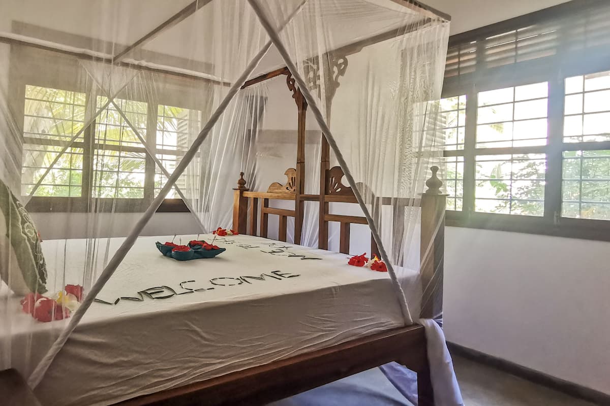 A room with a bed under a canopy, a wooden chair, and a window with a view of greenery.