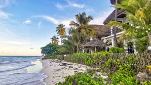 A beach with a wooden fence, palm trees, and a building with a thatched roof. - Villa Malaika (Jambiani)