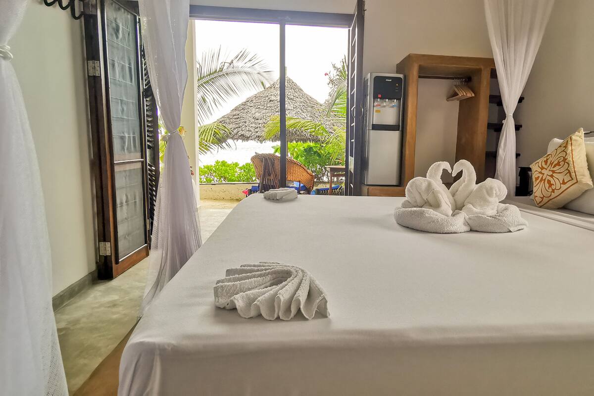 A hotel room with a bed, white curtains, a water dispenser, and a view of a tropical outdoor area.