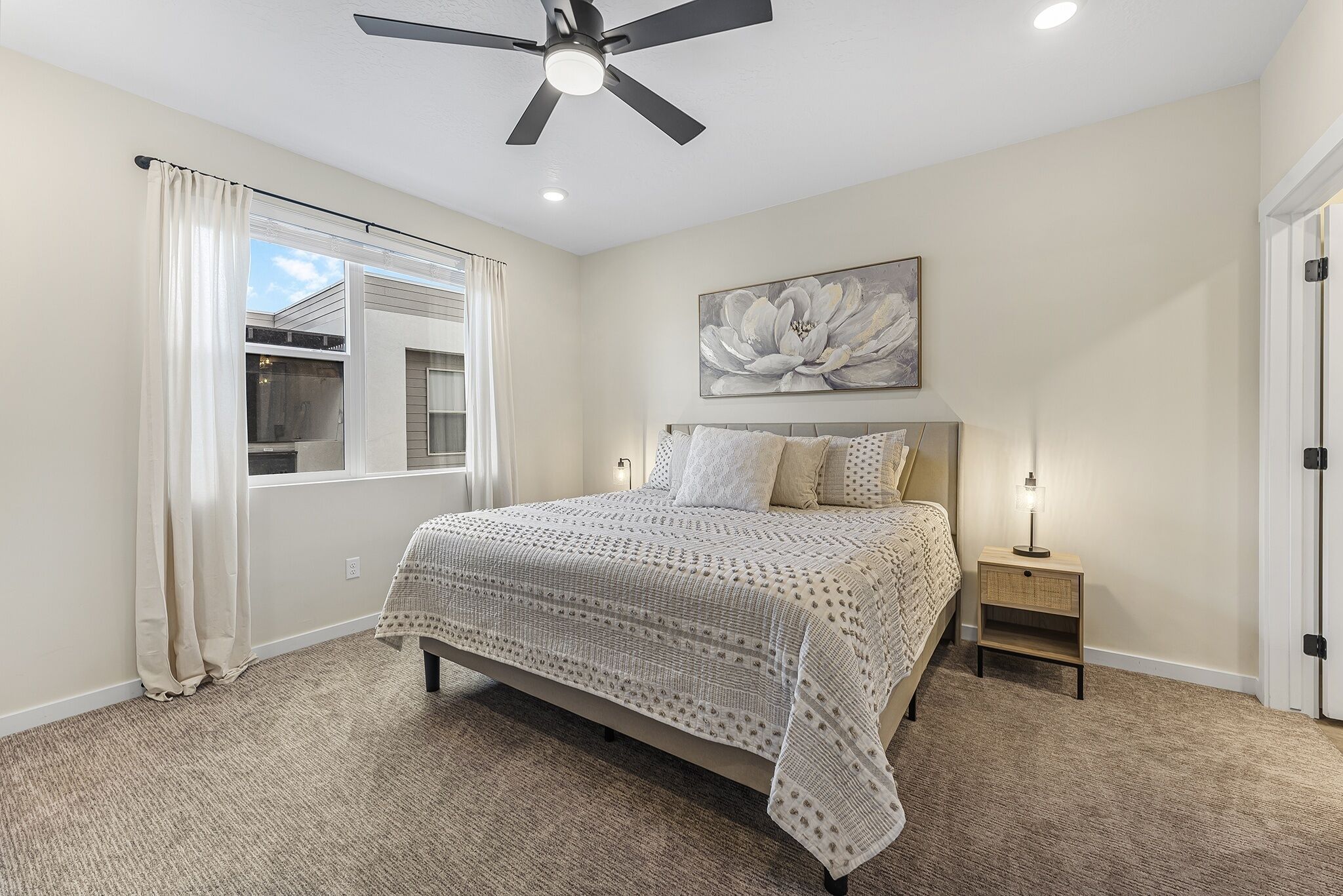 A bedroom with a bed, a ceiling fan, a window with curtains, a nightstand with a lamp, and a wall-mounted artwork.