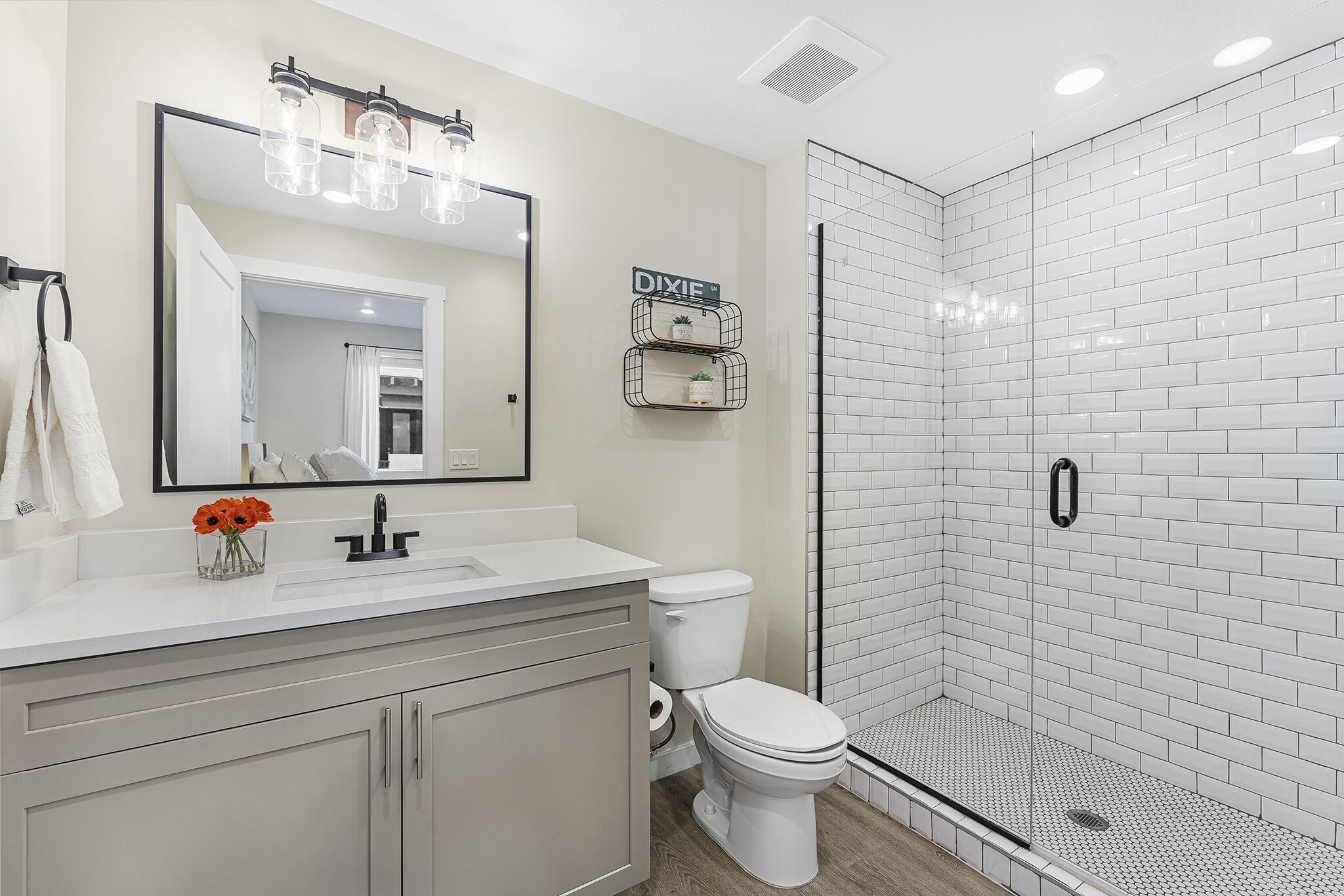 Un baño moderno con un lavamanos blanco, inodoro, ducha con azulejos blancos y un espejo con luces arriba.