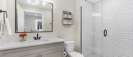 Un baño moderno con un lavamanos blanco, inodoro, ducha con azulejos blancos y un espejo con luces arriba.