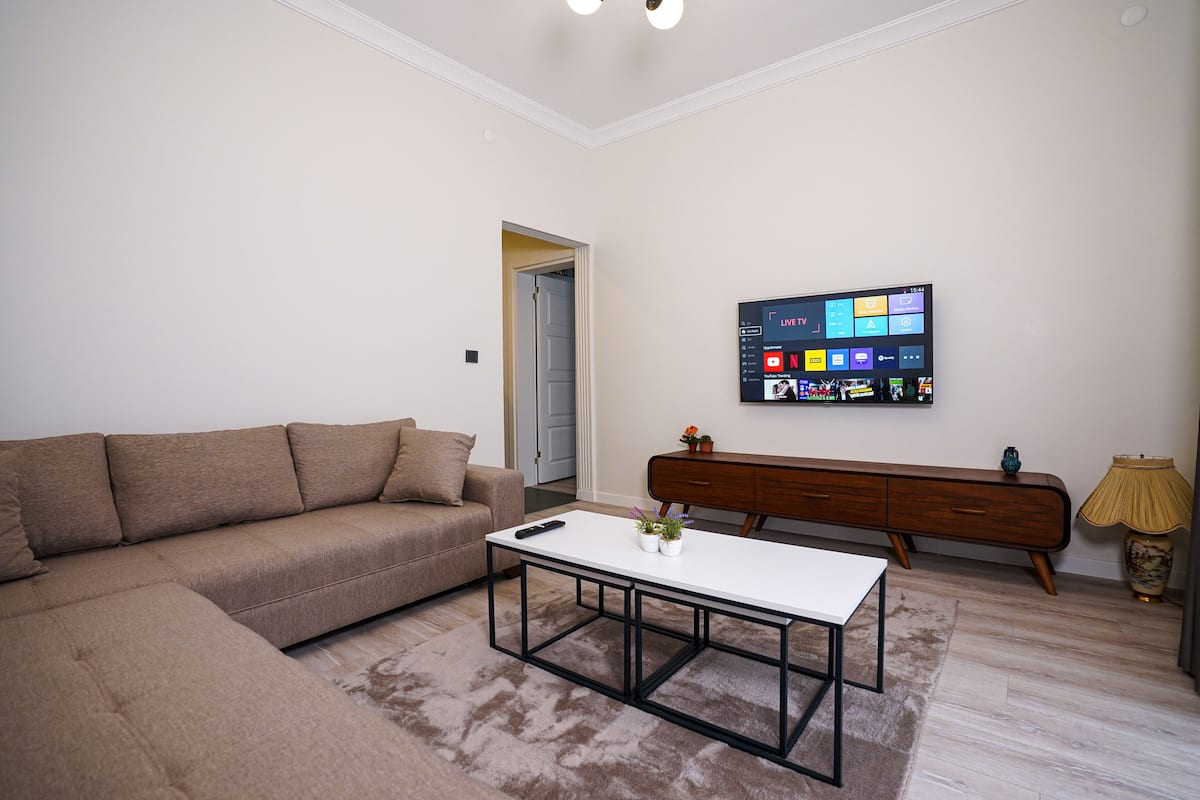 A modern living room with a sectional sofa, a coffee table, a TV mounted on the wall, and a wooden sideboard.