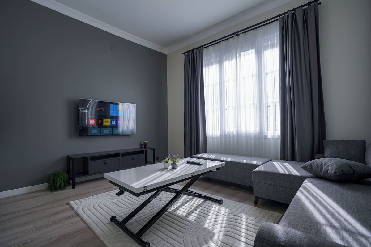 A modern living room with a flat-screen TV, grey sofa, and a large window with curtains.