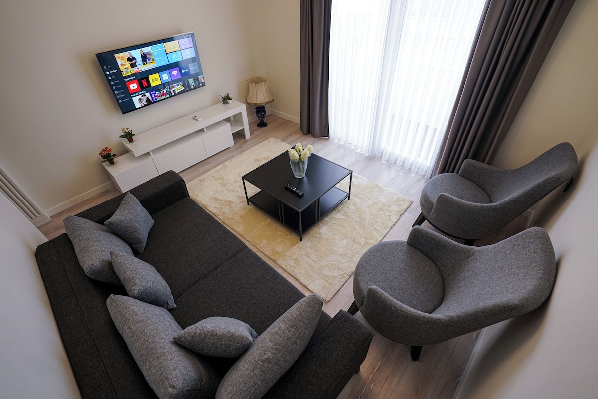 A modern living room with a black sofa, two grey armchairs, a black coffee table, and a flat-screen TV mounted on the wall.