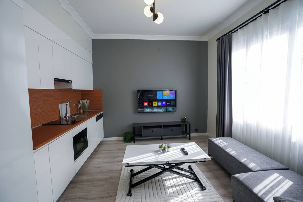 A modern living room with a sofa, a coffee table, a TV, and a kitchen area with cabinets and a countertop.