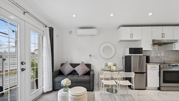 Eine moderne Wohnung mit Balkon, einem grauen Sofa mit Kissen, einem runden Esstisch mit Stühlen, einer Küche mit Edelstahlgeräten und einem weißen Klimagerät.