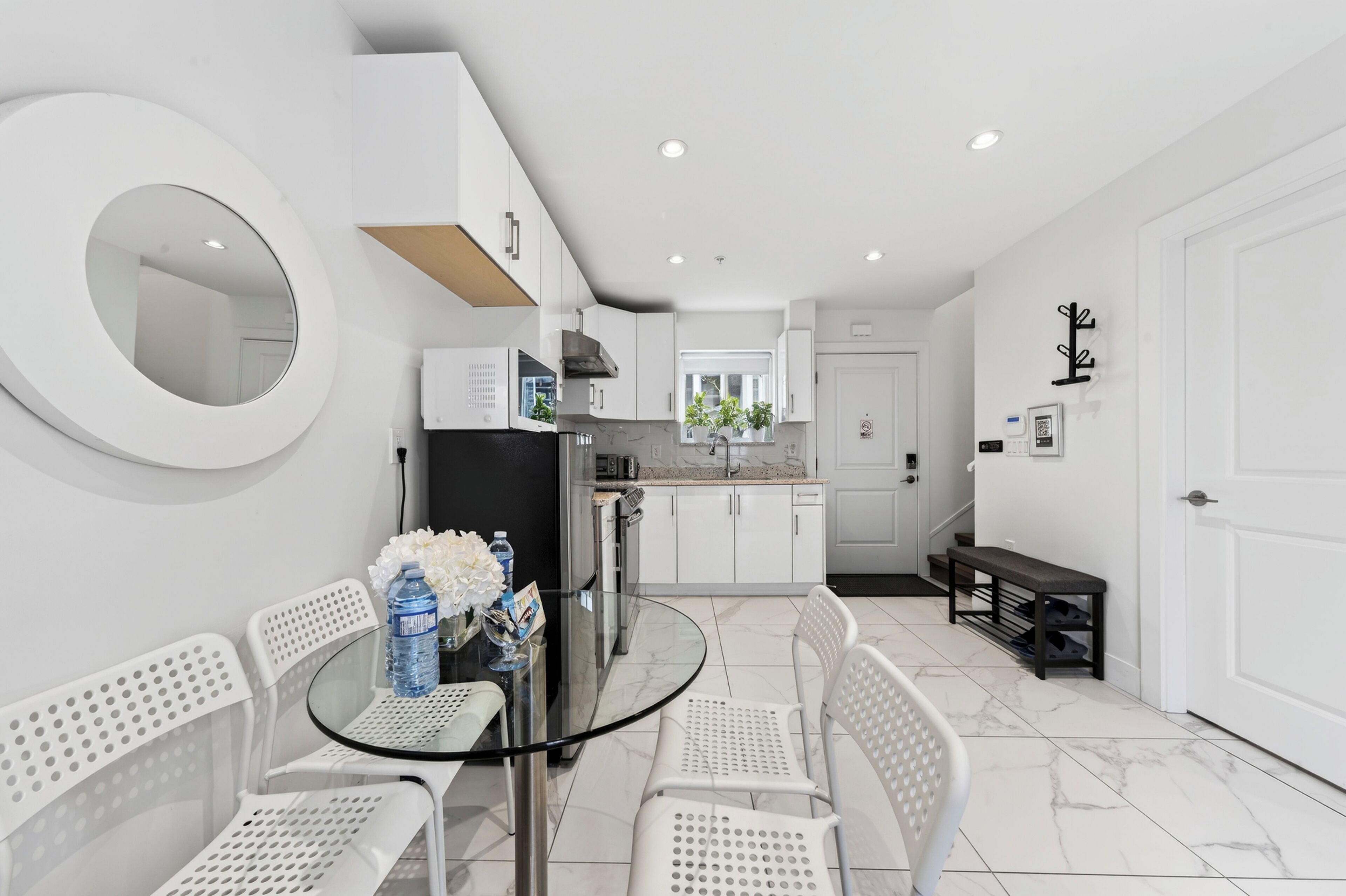 A modern kitchen with white cabinets, a round mirror, and a glass dining table with white chairs.