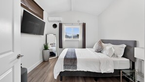 Ein modernes Schlafzimmer mit Bett, Fernseher, einem Fenster mit Vorhängen, einer Pflanze und einem Spiegel.