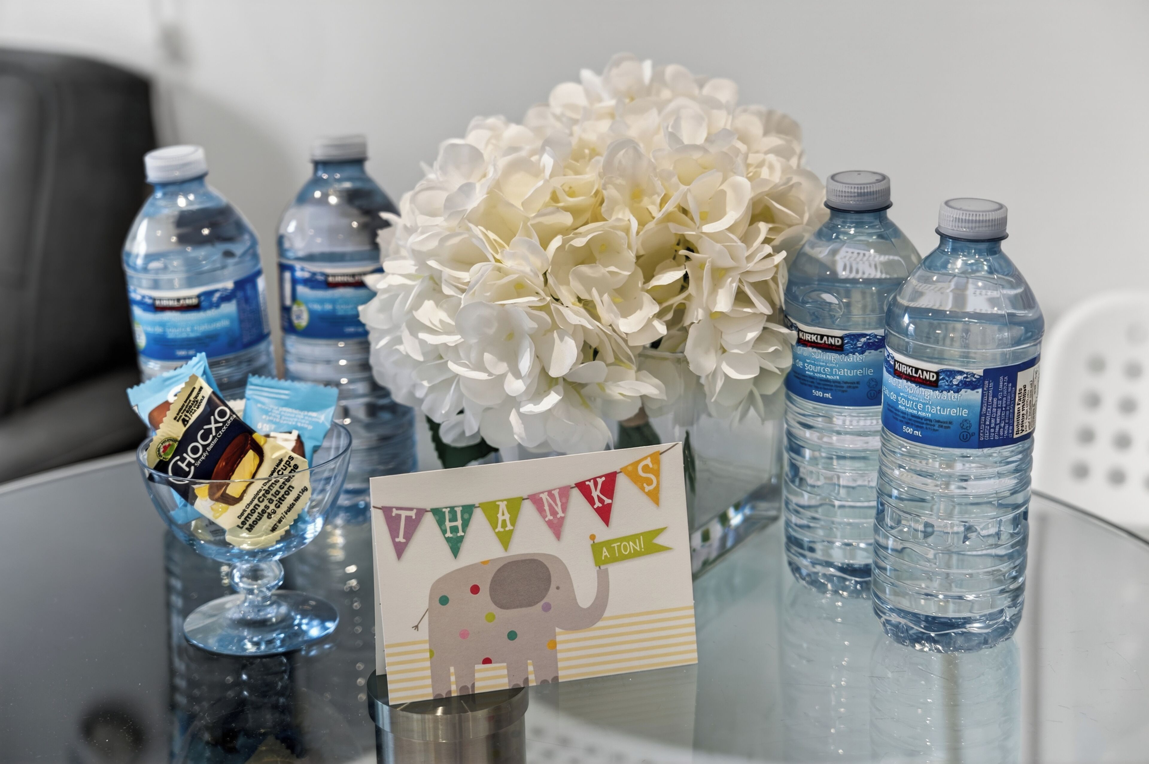 Ein Tisch mit Wasserflaschen, einer Schale mit Snacks, einer Dankeskarte und einer weißen Blumenkomposition.