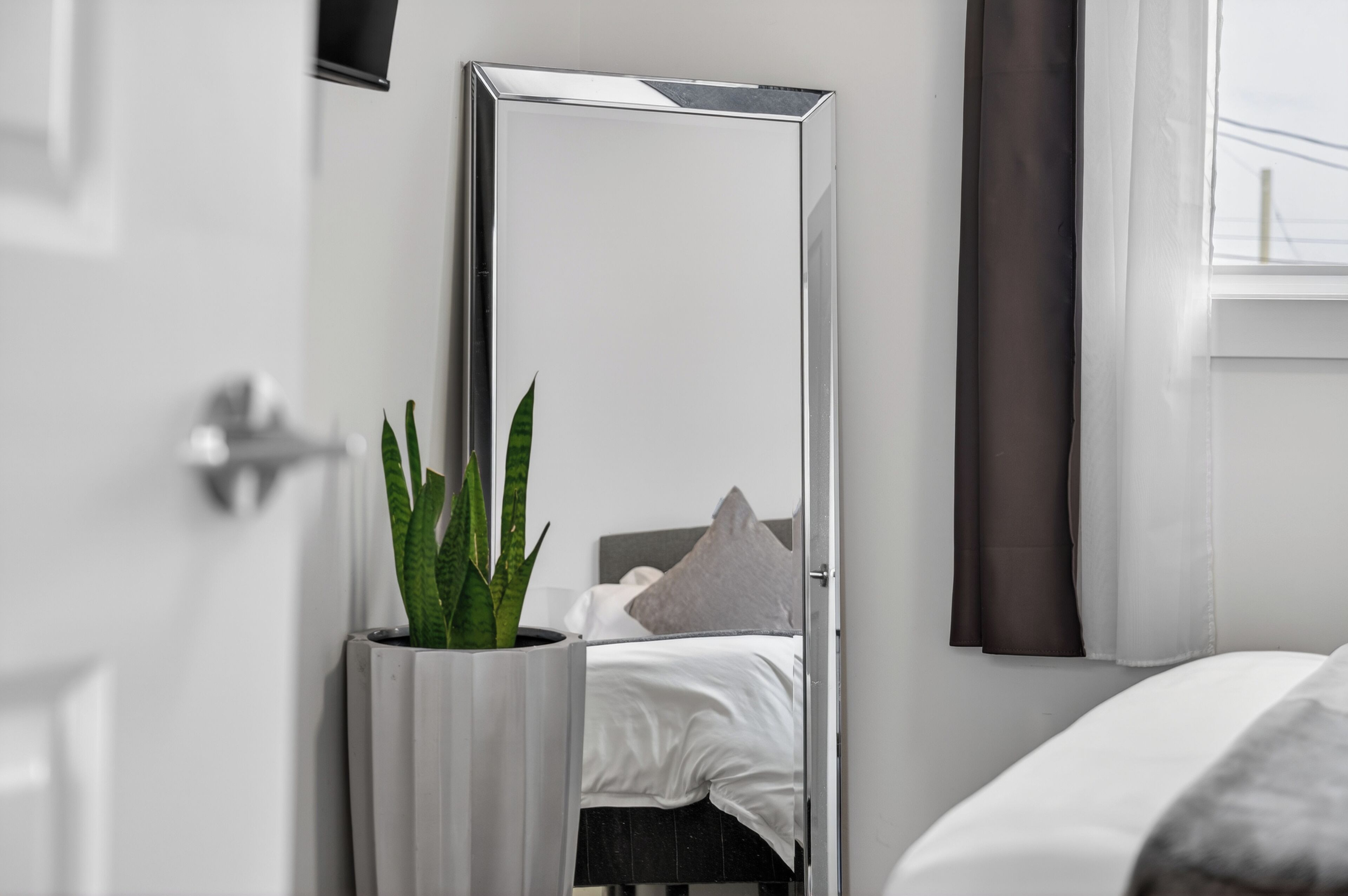 Une chambre d’hôtel moderne avec un grand miroir, une plante en pot, un lit avec une literie blanche et une fenêtre avec des rideaux.