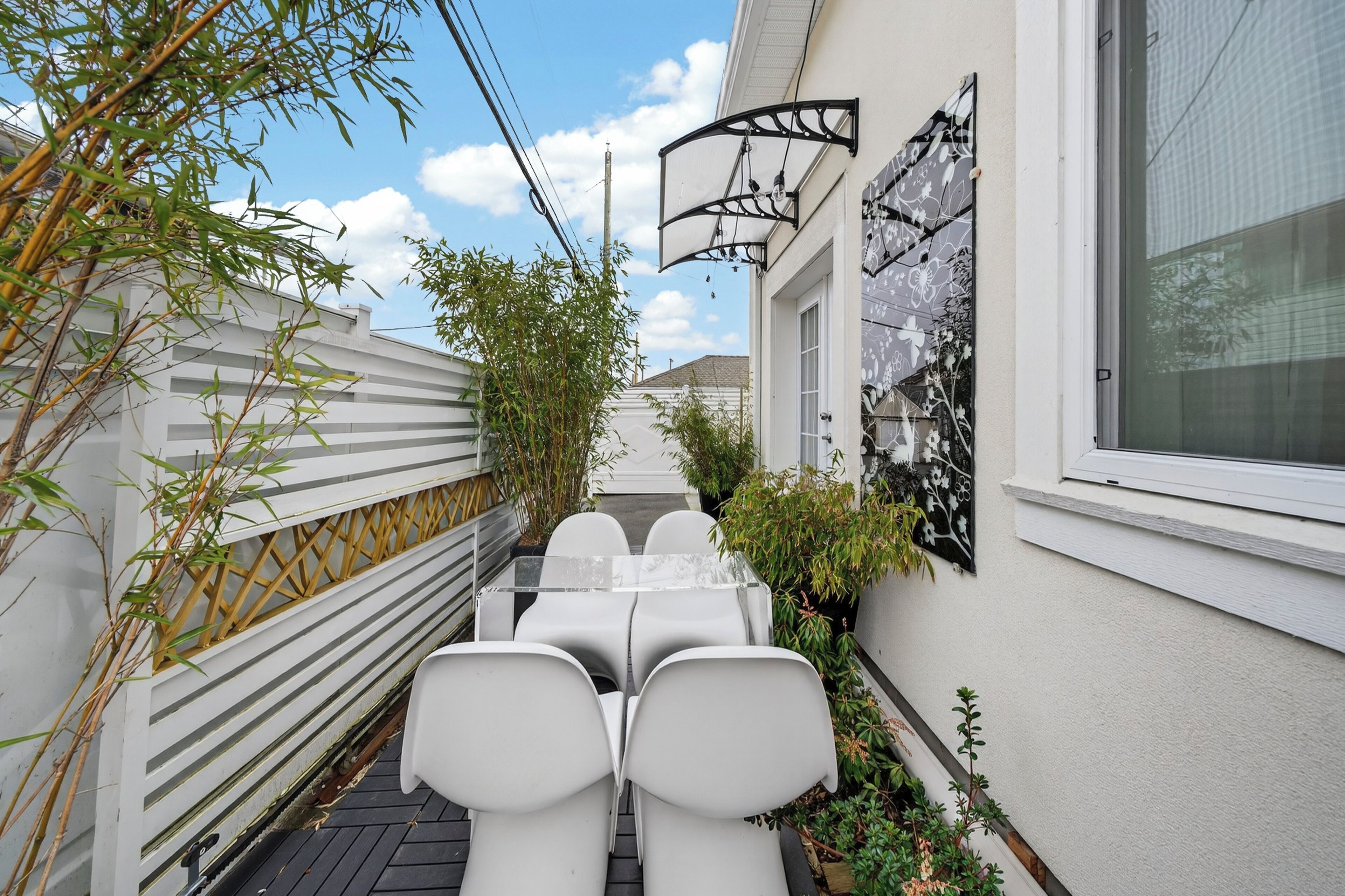 A narrow outdoor patio with a glass-top table and white chairs, surrounded by bamboo plants and a white fence.