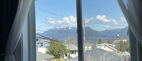 A view from a window with white curtains, overlooking a residential area with houses and a mountain range in the background.