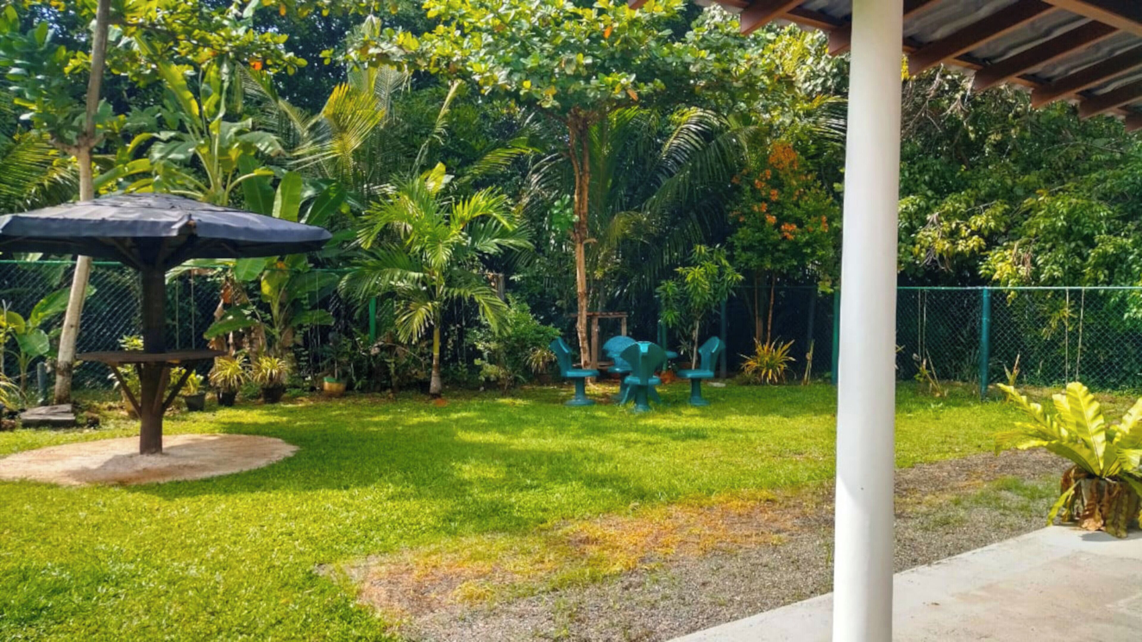 A covered outdoor seating area with a table and chairs, surrounded by lush greenery and a fence.