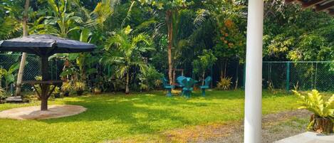 A covered outdoor seating area with a table and chairs, surrounded by lush greenery and a fence.