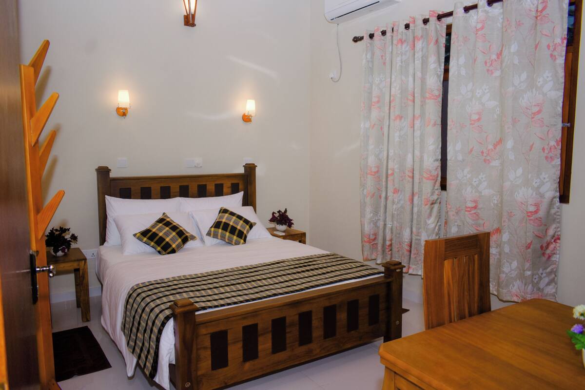 Un dormitorio con una cama de madera, dos mesitas de noche, un buró, una silla de madera, una mesa de madera y una ventana con cortinas florales.