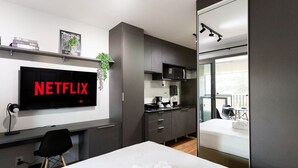 A modern hotel room with a large bed, a desk with a computer, a TV displaying 'NETFLIX', a kitchenette, and a balcony with a view. - Studios Hub Vila Madalena 2.0 (São Paulo)