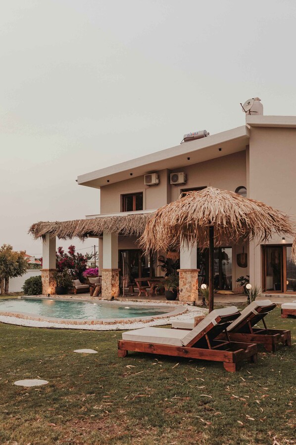A modern house with a swimming pool, outdoor seating, and a thatched roof structure.