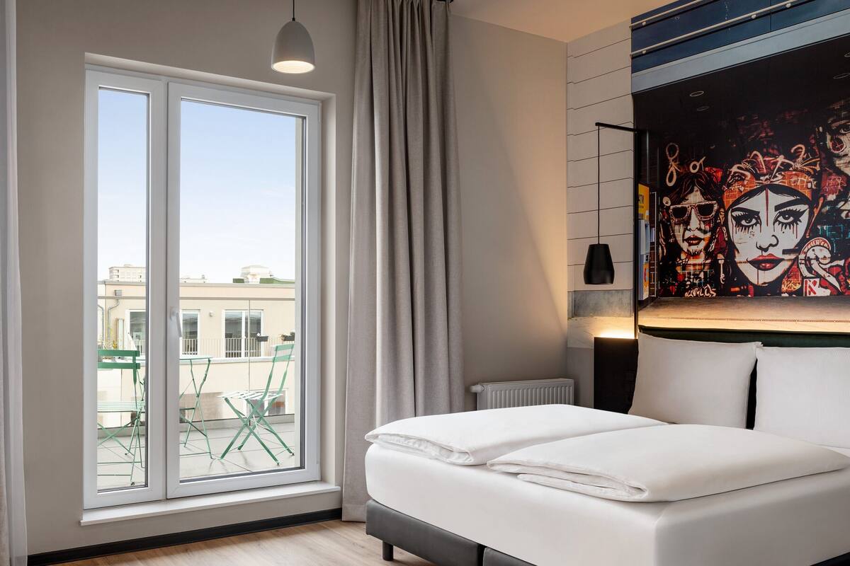 A hotel room with a large window, a bed, a green chair, and a wall mural.