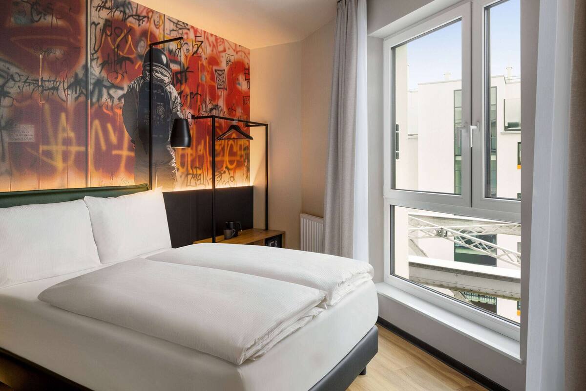 A modern hotel room with a large bed, a view of the city through a window, and a graffiti-covered wall.