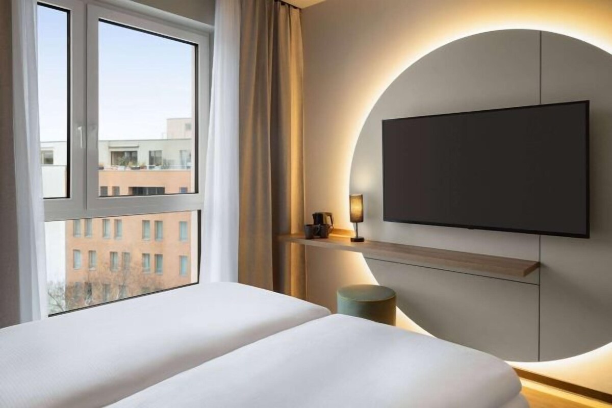 A modern hotel room with a large bed, a TV mounted on the wall, and a view of buildings outside the window.