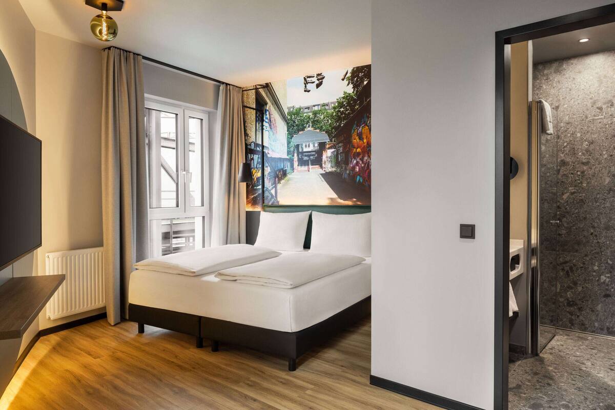 A modern hotel room with a large bed, a view of a street through a window, and a shower area.