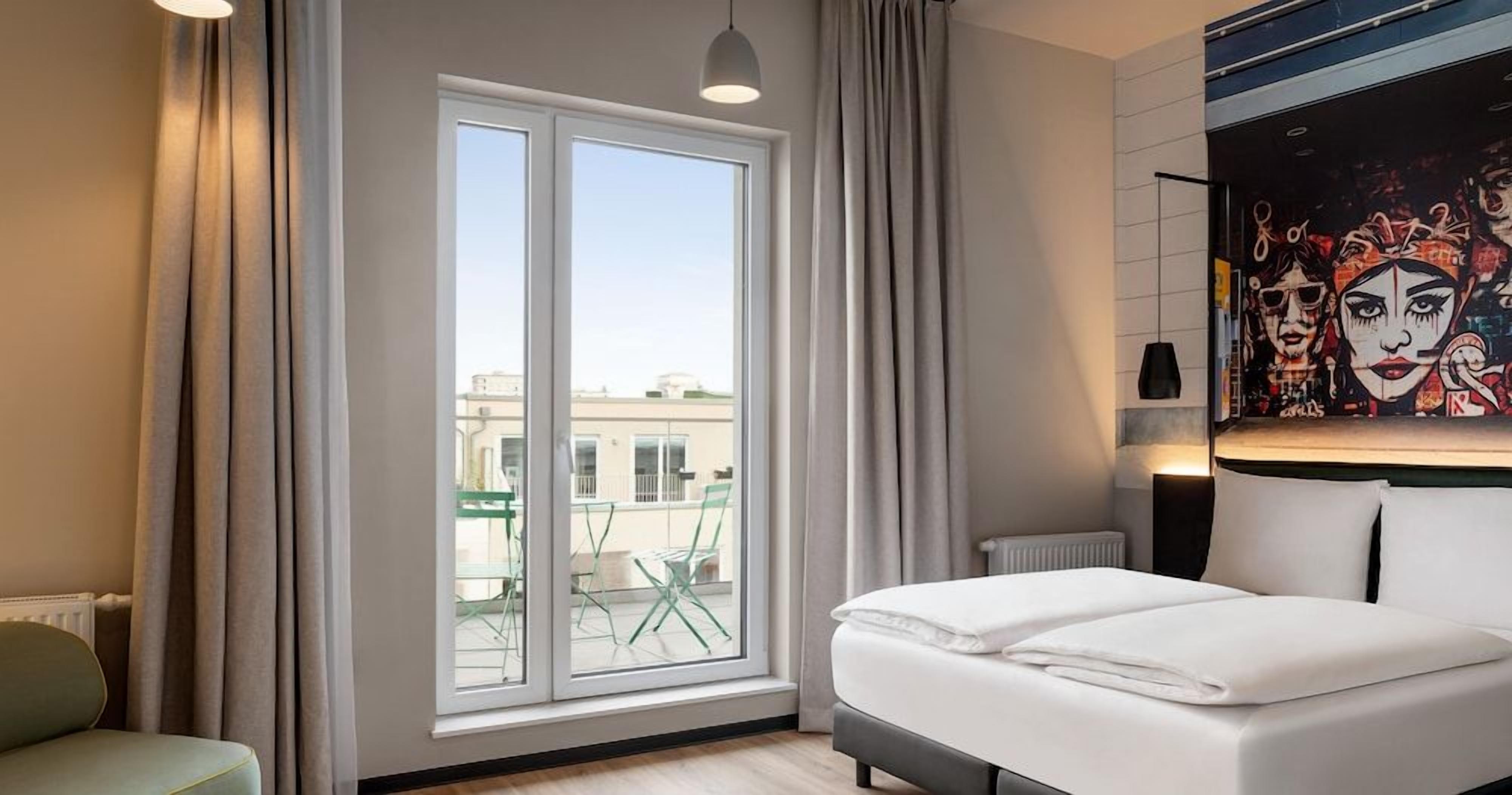 A hotel room with a large window, a bed, a chair, and a painting on the wall.