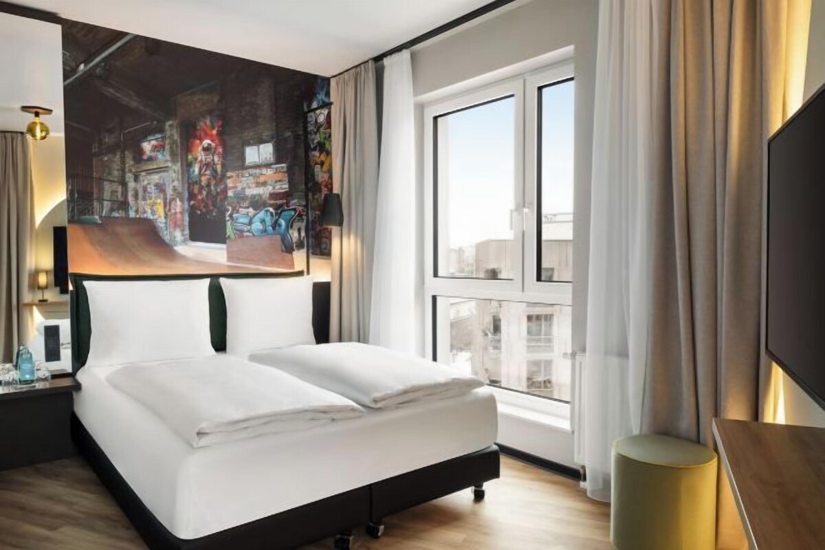 A modern hotel room with a large bed, a cityscape mural on the wall, a large window with curtains, and a flat-screen TV.