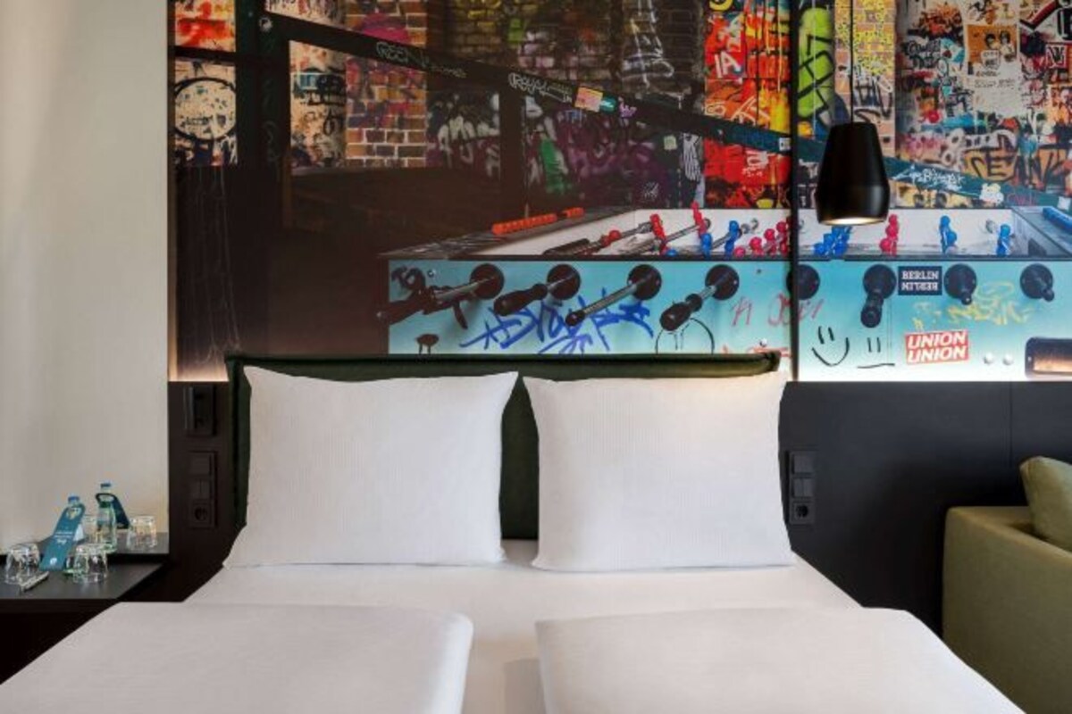 A hotel room with a bed, pillows, a nightstand with a lamp, and a wall with graffiti art.