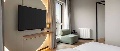 A modern hotel room with a large bed, a flat-screen TV mounted on the wall, a small seating area, and a window with curtains.
