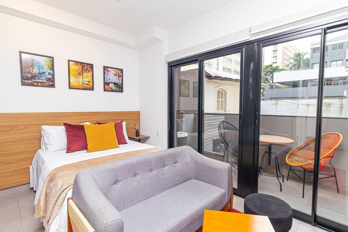 A hotel room with a bed, a grey sofa, a small table, a balcony with orange chairs, and three paintings on the wall.