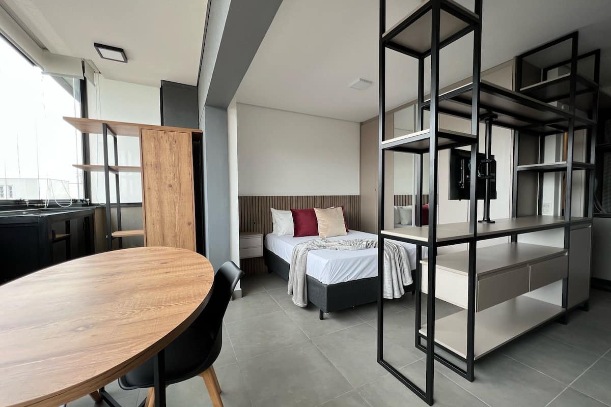 A modern hotel room with a wooden dining table, a bed with a headboard, a built-in wardrobe, and a large window.