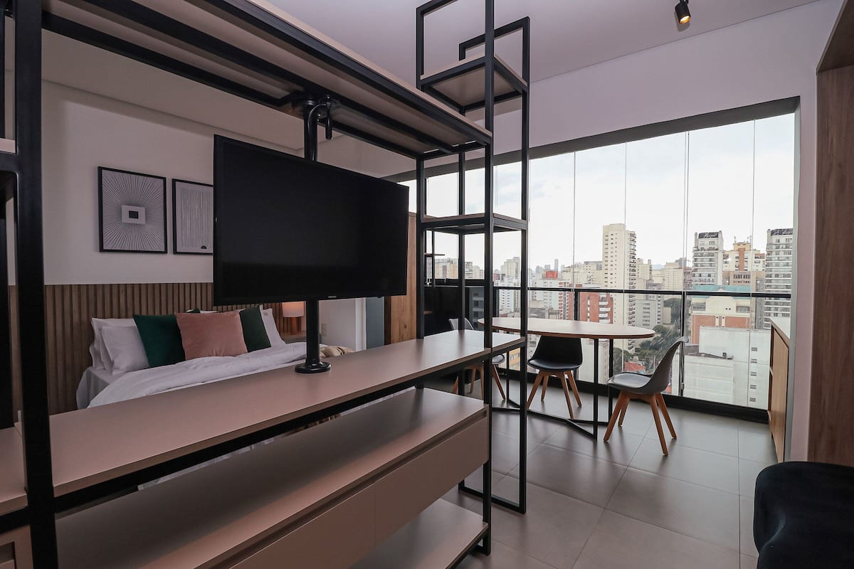 A modern hotel room with a bed, a desk, a TV, and a balcony with a city view.