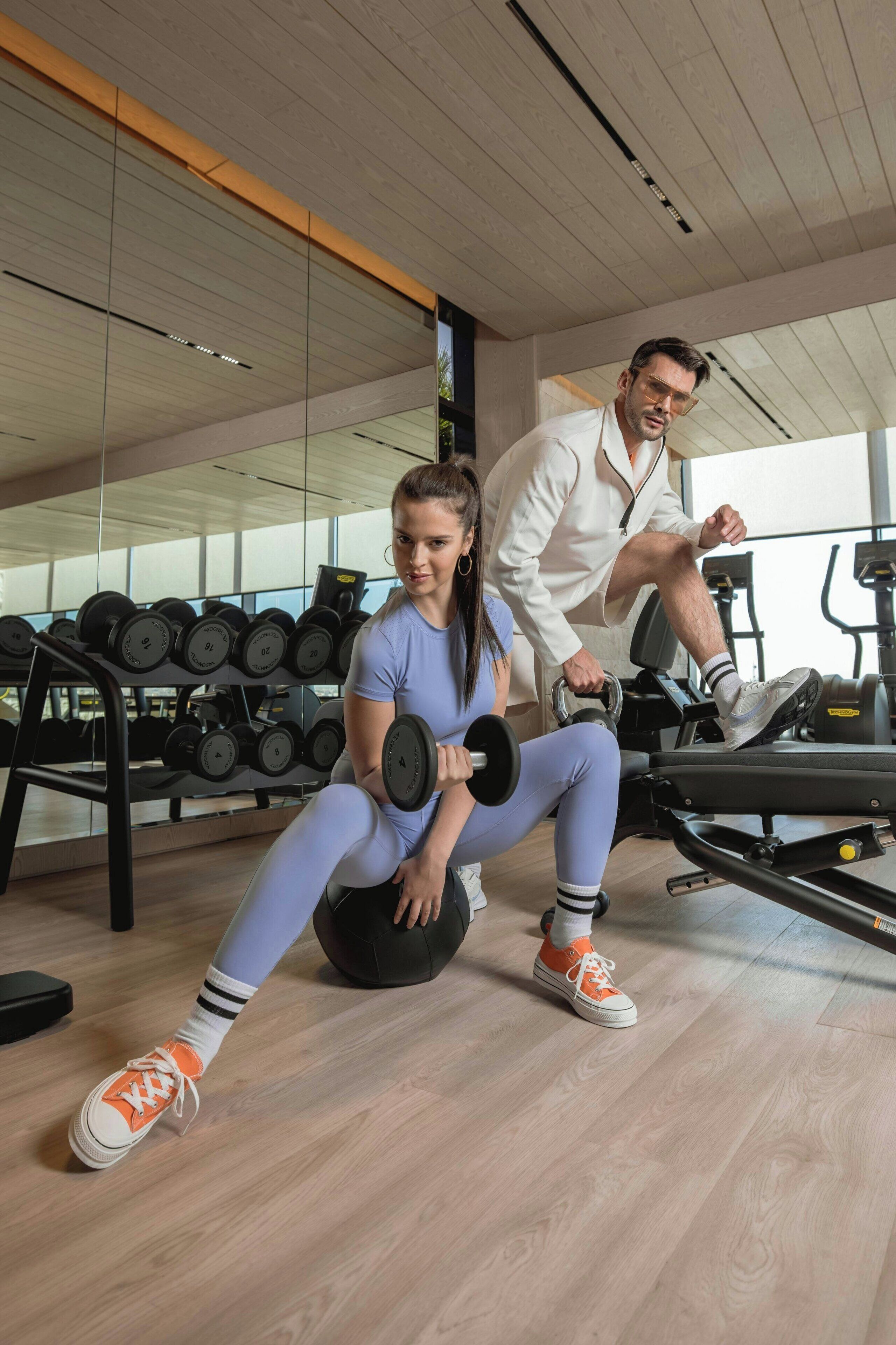 A gym with exercise equipment, a woman and a man performing exercises, and a rack of dumbbells.
