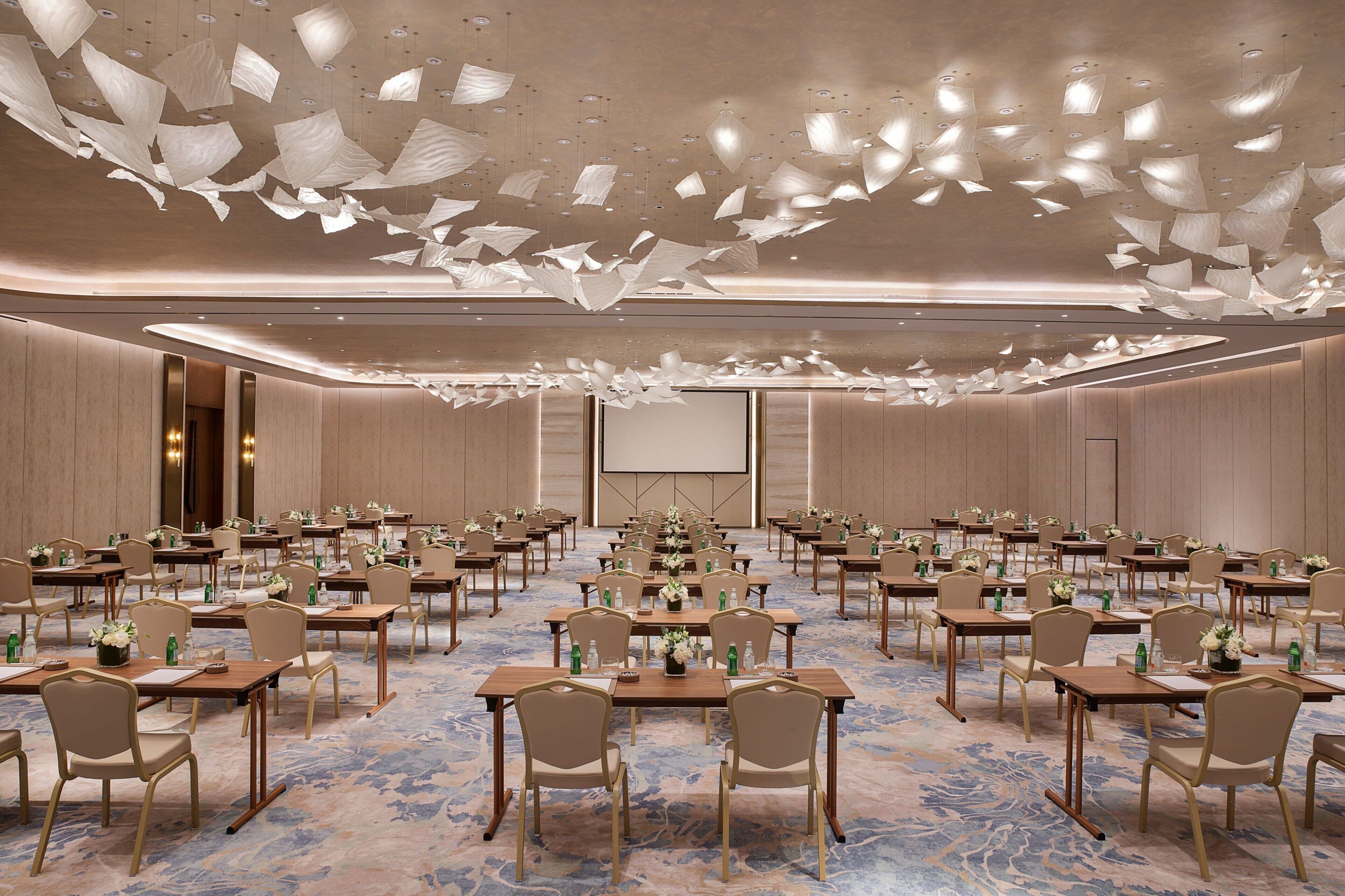 Una sala conferenze con file di tavoli e sedie, un grande schermo e un soffitto con lampade a sospensione.