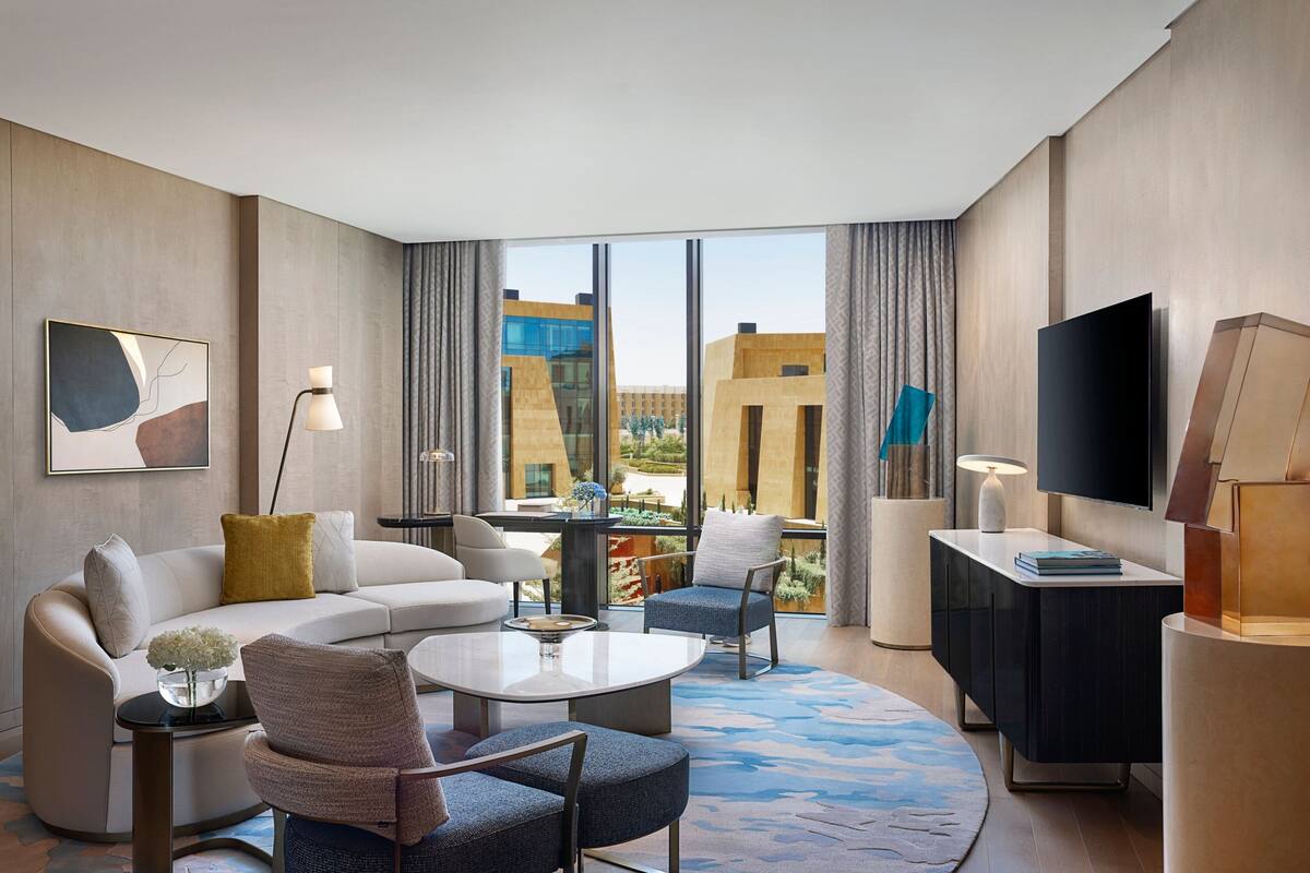 A modern hotel room with a large window, a flat-screen TV, a sofa, a coffee table, and a dining table.