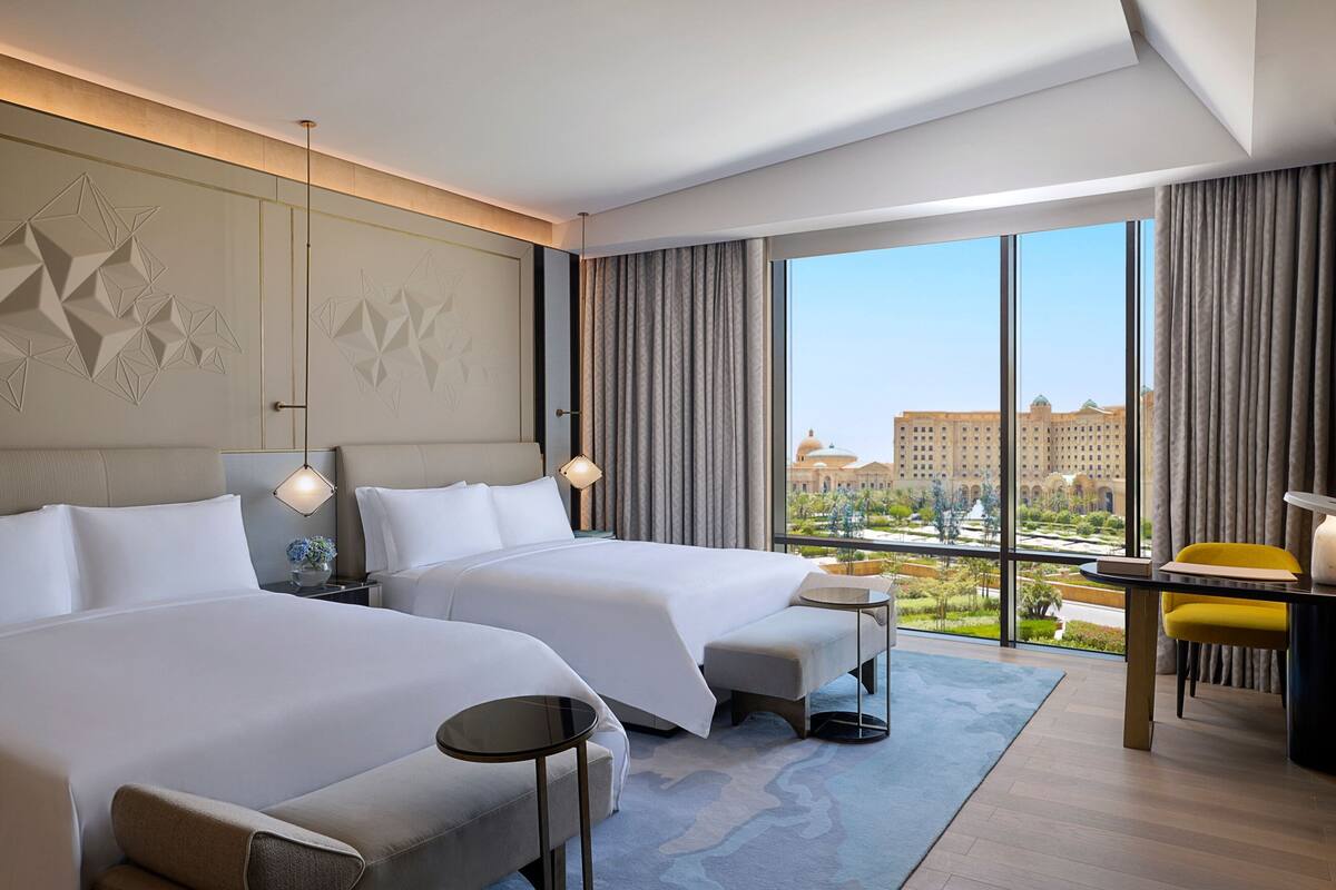 A hotel room with two beds, a desk, and a view of a cityscape.